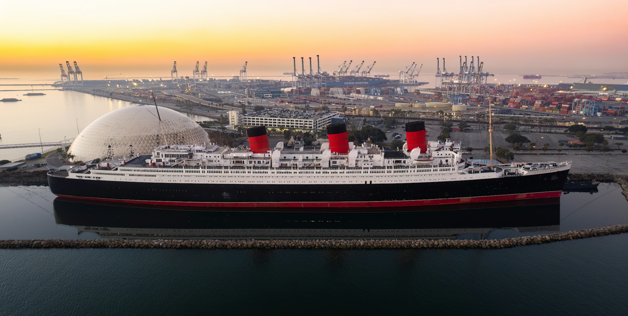 A large retired ocean liner docked with a port and sunset in the background.