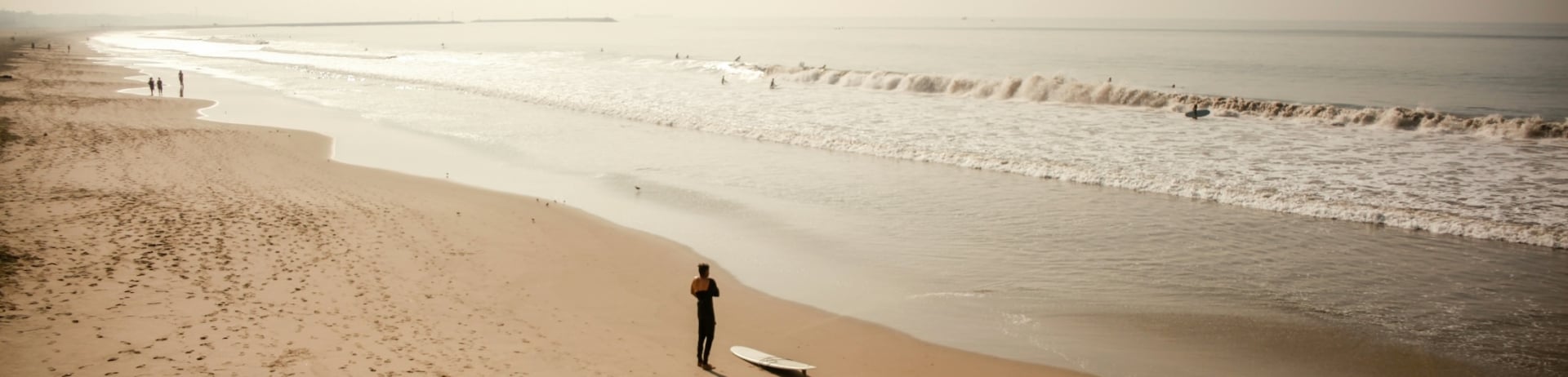 A man zips up his wetsuit next to his surf board on the beach while other surfers are in the waves.