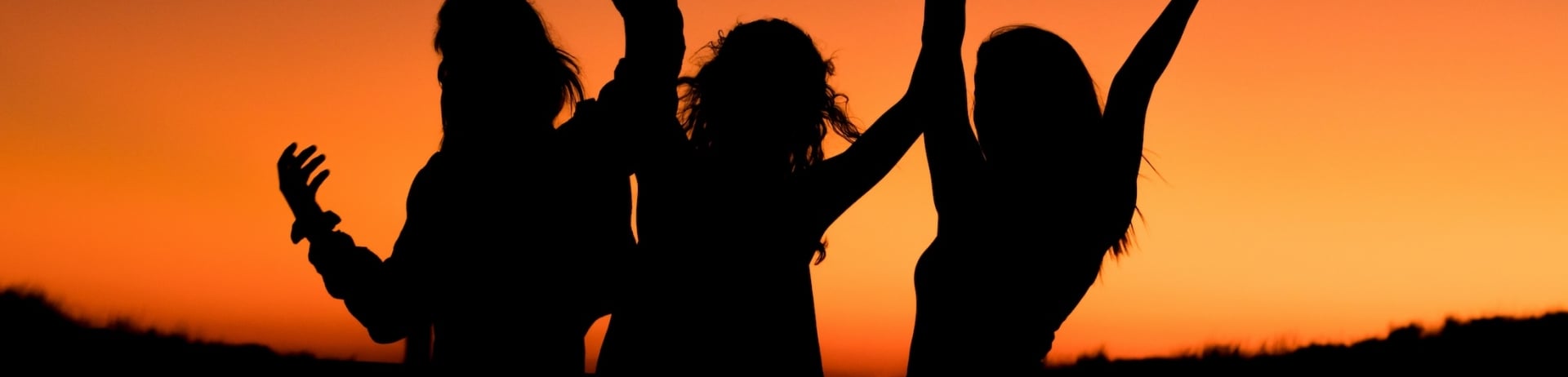 The silhouette of three women with their hands in the air against an orange sunset.