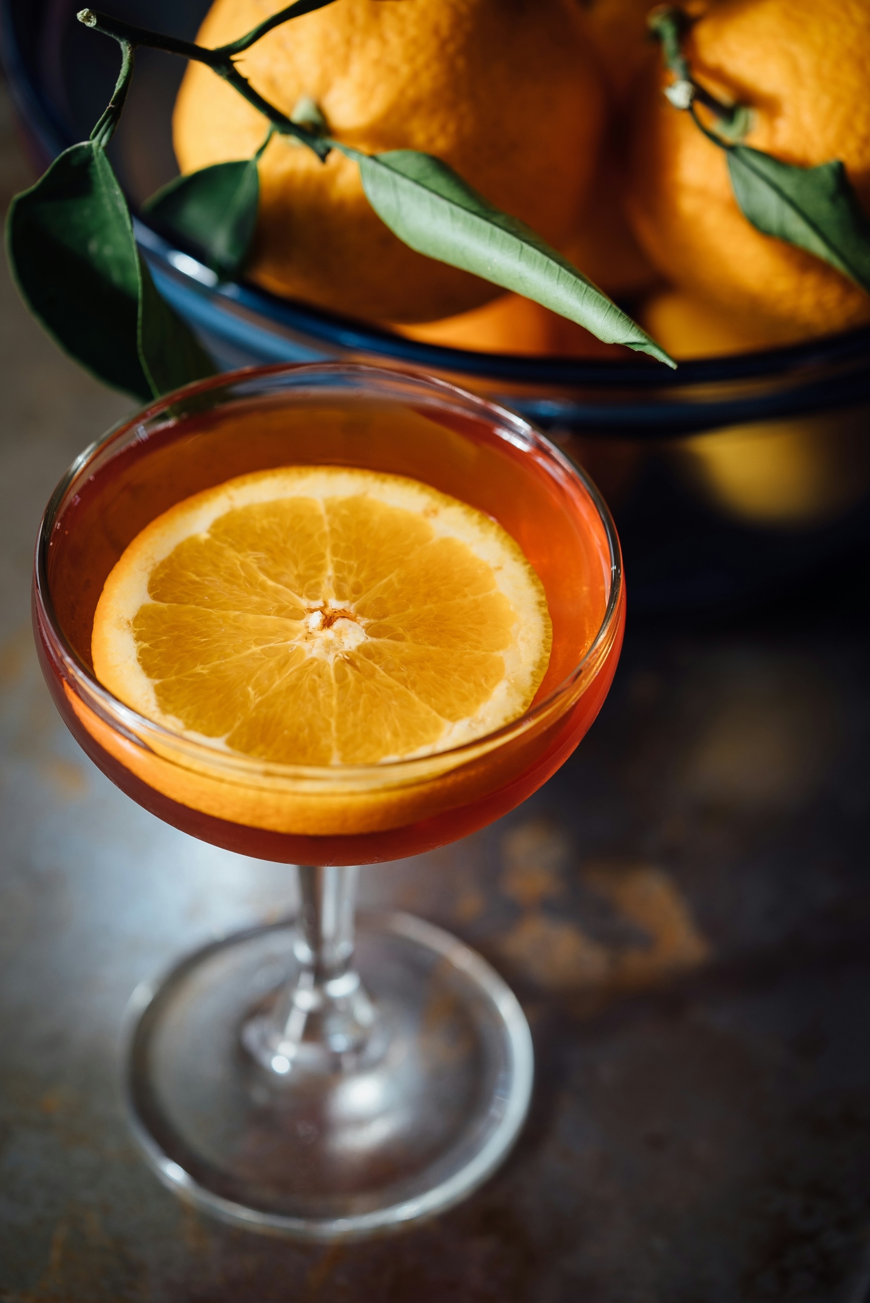A red cocktail in a coupe glass garnished with a round slice of orange. There is a bowl of oranges in the background.